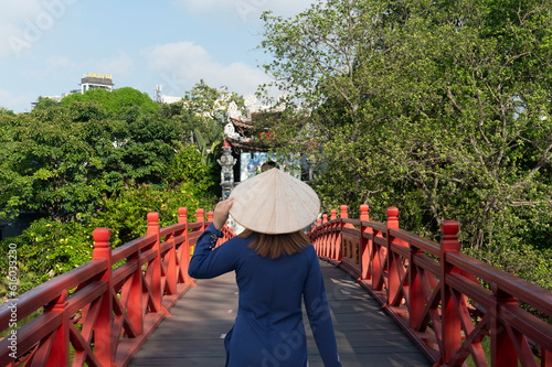 Asian woman is wearing Ao Dai traditional Vietnamese dress and traveling at Red Bridge- The Huc Bridge in Hoan Kiem Lake, this is a lake in the historical center of Hanoi, the capital city of Vietnam photo