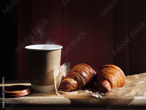 Morning coffee cup and croissants on a wooden table in natural sunlight, dark blur background. Coffee takeaway with steam of hot drink and two fresh french croissants on paper. Breakfast meal in city.