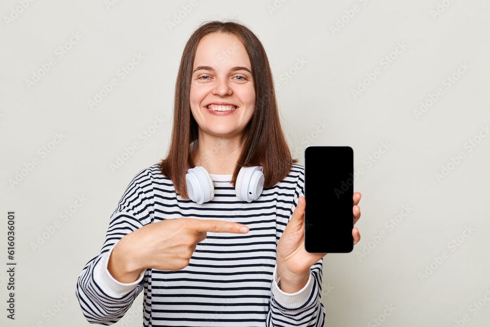 Fototapeta premium Attractive cheerful woman wearing striped shirt standing with headphones over neck isolated over gray background pointing at smartphone with empty display copy space for advertisement