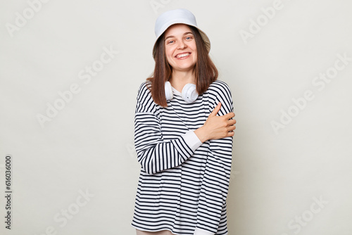 Принти на полотні Pleased woman wearing striped shirt and panama standing with headphones over neck isolated over gray background hugging herselfexpressing self loving