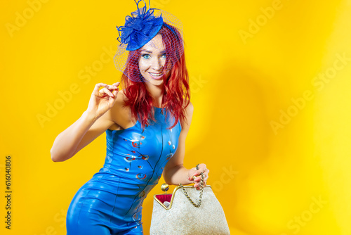 Expressive Caucasian Blond Woman in Blue Artistic Dance Suit Posing In Shawl Hat With Extravagant Hand-Bag Against Yellow Background.