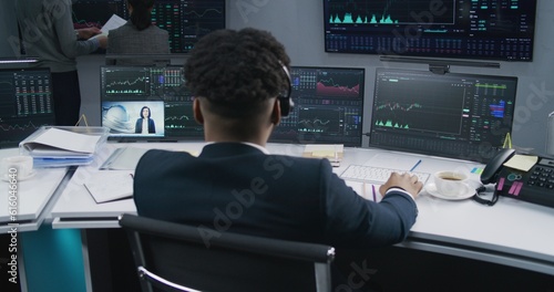African American trader works at computer with displayed real-time stocks. Coworkers analyze exchange market charts on big screens at background. Trading and investment concept. Back view. Dolly shot.
