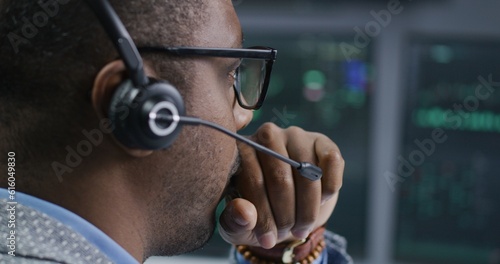 African American financial analyst in headset working in bank office. Businessman looks at computer screen  monitors real-time stocks  exchange market charts. Investment and analytics. Close up.