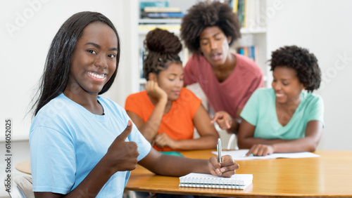 Beautiful black female student at desk showing thumb up with group of learning african american students