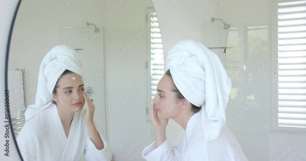 Caucasian woman with towel on head applying cream on face in bathroom in slow motion Caucasian woman with towel on head applying cream on face in bathroom in slow motion