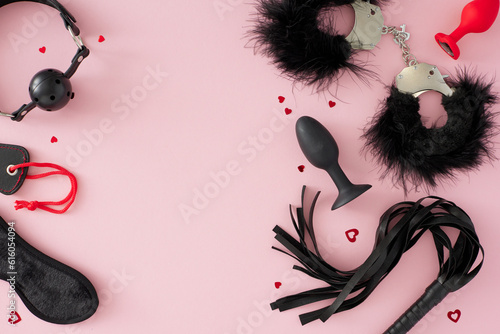 Array of pleasure toys for BDSM adventures. Top view photo of feather handcuffs, leather whip, ball gag, anal plugs, red hearts on pastel pink background with empty space for text or advert