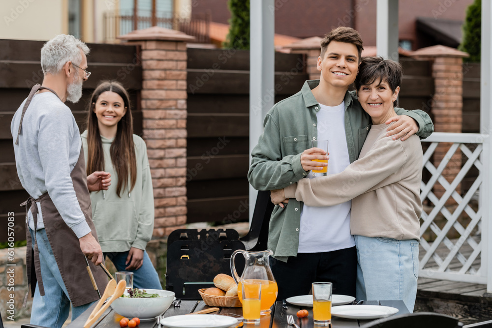 happy parents day, modern parenting, cheerful middle aged mother hugging young adult son holding glass of orange juice, father and daughter preparing food on bbq grill, summer, backyard, celebration happy parents day, modern parenting, cheerful middle aged mother hugging young adult son holding glass of orange juice, father and daughter preparing food on bbq grill, summer, backyard, celebration