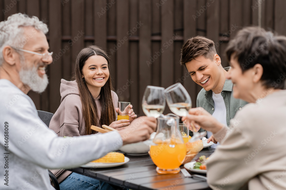 Smiling siblings sitting near tasty bbq food and blurred middle aged parents toasting with wine during parents day celebration at backyard, family love and unity concept, tradition and celebration Smiling siblings sitting near tasty bbq food and blurred middle aged parents toasting with wine during parents day celebration at backyard, family love and unity concept, tradition and celebration