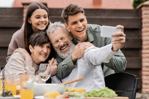 Cheerful kids hugging middle aged parents while taking selfie on smartphone together during barbeque party and parents day celebration at backyard in june  happy parents day concept
