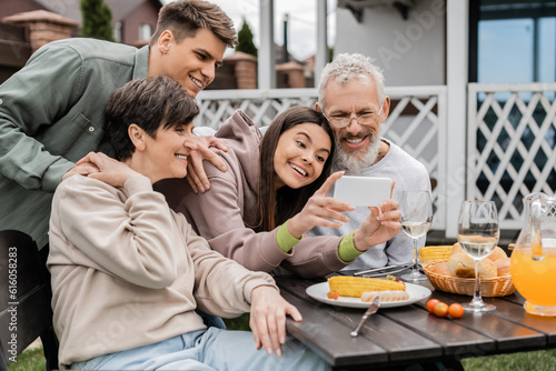 Positive teenage girl using smartphone near mature parents and father during bbq party with summer food and parents day celebration at backyard in june  happy parents day concept  special occasion