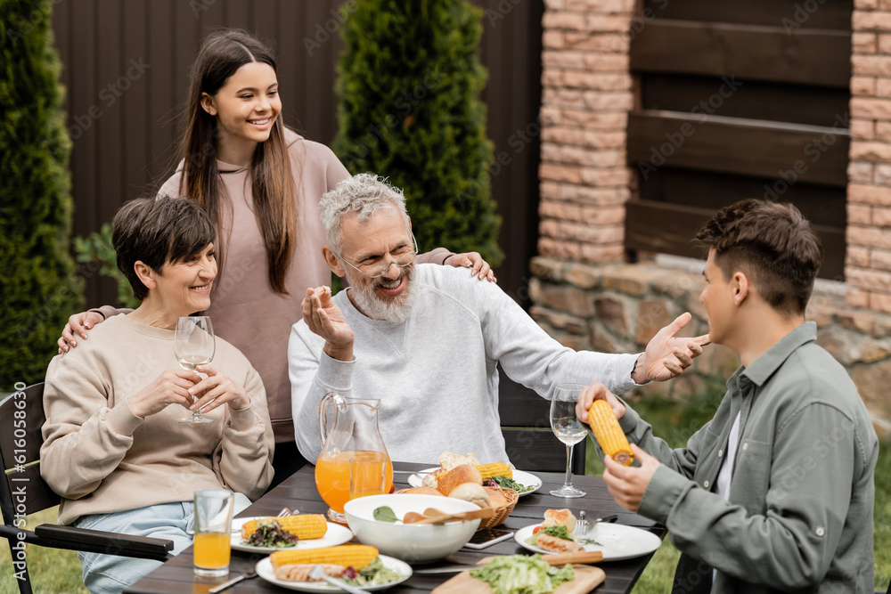 Cheerful middle aged man talking to young soon while sitting near family and summer food during bbq party and parents day celebration at backyard, special day for parents concept