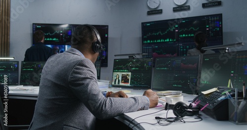 African American trader with pen in hand works at computer with displayed real-time stocks. Colleagues analyze exchange market charts on big screens at background. Concept of trading and investment.