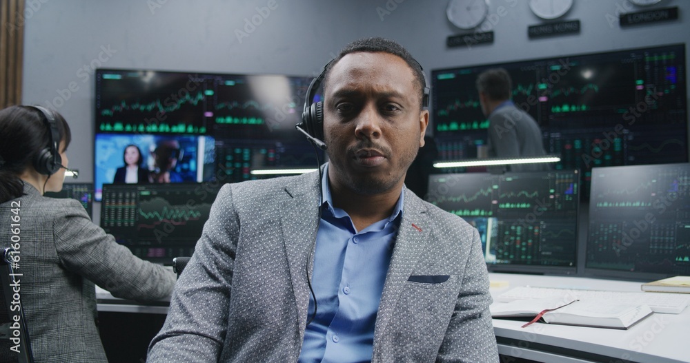 Obraz premium Tired African American financial analyst sitting at workplace takes off glasses and looks at camera. Colleagues analyze exchange market charts on screens at background. Investment concept. Portrait.