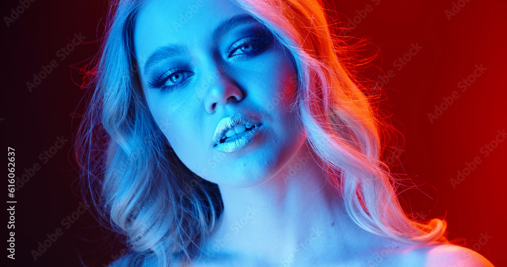 Sensual blonde caucasian girl with glowing makeup giving a tempting look in neon cyberpunk lights - nightlife concept close up portrait shot 