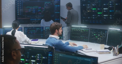 Technical support specialists sit at computers with displayed blockchain network map and server data. Big data scientists work in monitoring room. Big screens on the wall. Concept of cyber security.