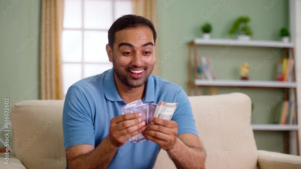 Happy excited indian man counting money or currency notes while sitting on sofa at home - concept of profit, won lottery and Financial success. Happy excited indian man counting money or currency notes while sitting on sofa at home - concept of profit, won lottery and Financial success.