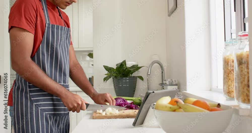 Biracial man in apron chopping vegetables and using tablet in kitchen, slow motion Biracial man in apron chopping vegetables and using tablet in kitchen, slow motion