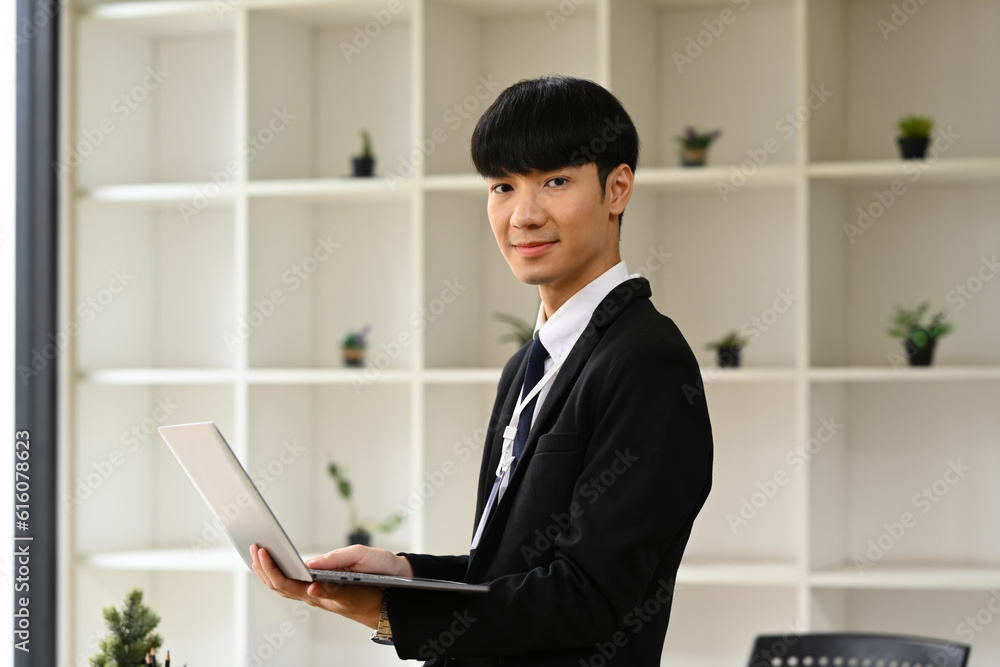 Portrait of young businessman in black suit holding laptop and looking confidently to camera