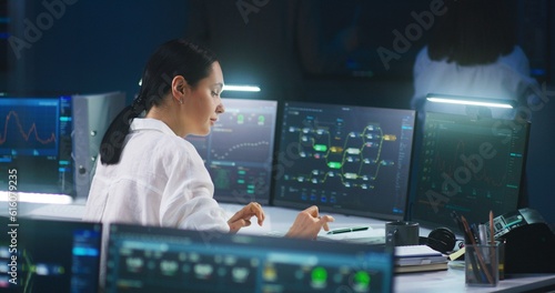 Female IT technical specialist works on computer with data server and blockchain network database in modern monitoring control room. Team of software engineers and big digital screens on background.