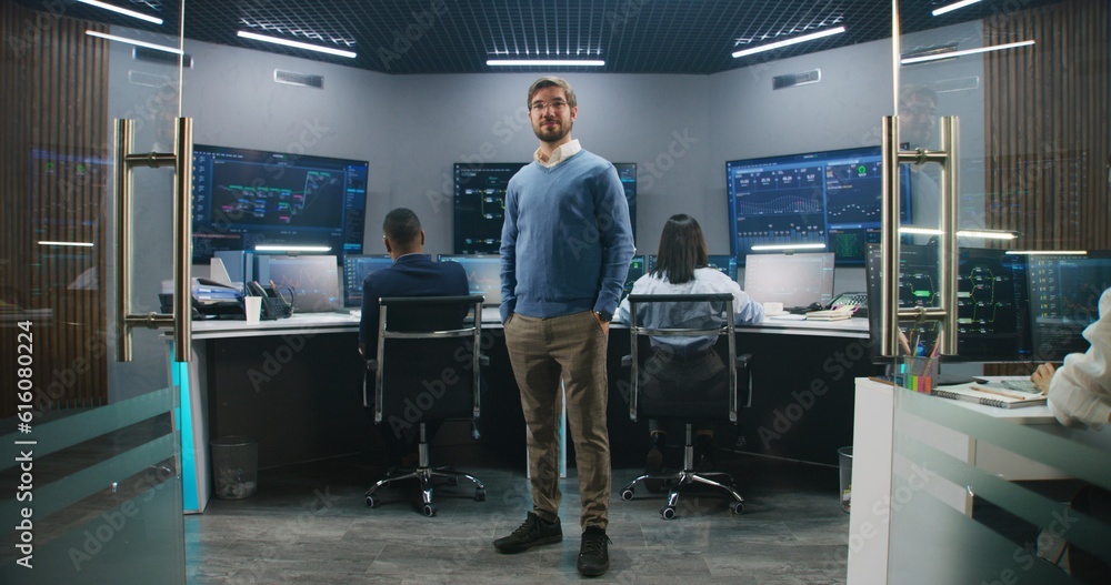 Adult IT scientist smiles, stands in monitoring room and looks at camera. Big data engineers work at computers at background. Multiple big screens with displayed real-time analysis charts. Dolly shot. Adult IT scientist smiles, stands in monitoring room and looks at camera. Big data engineers work at computers at background. Multiple big screens with displayed real-time analysis charts. Dolly shot.