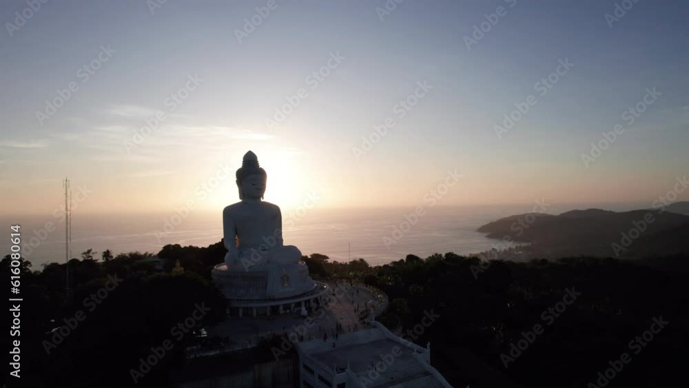 Big Buddha at sunset view from a drone. Phuket. People climb the steps to the statue. The green hills of the island are all around. In the distance, the bright sun goes over the sea. View from above Big Buddha at sunset view from a drone. Phuket. People climb the steps to the statue. The green hills of the island are all around. In the distance, the bright sun goes over the sea. View from above