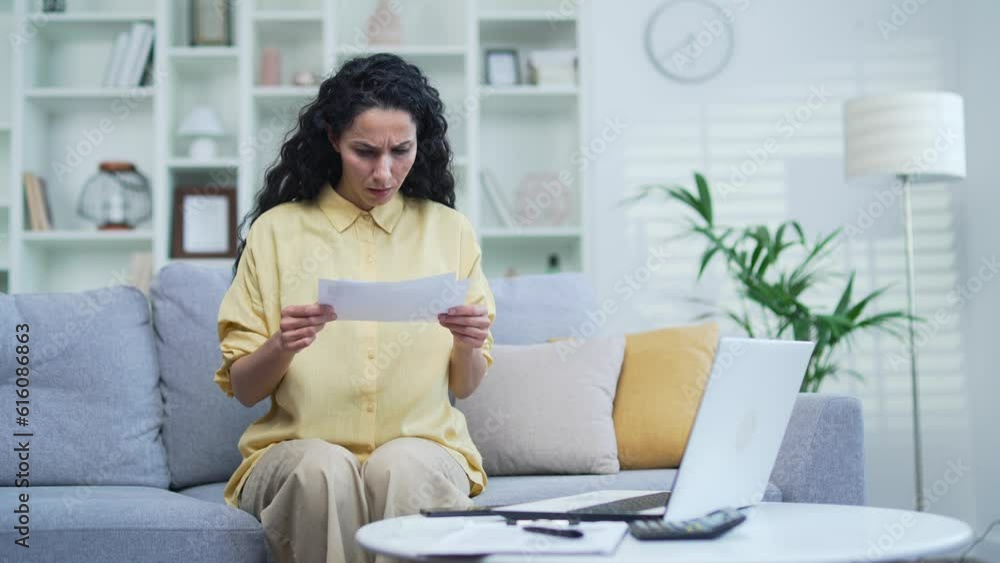 Frustrated shocked woman looking at large utility bills sitting on the sofa in the room at home. Upset female holding a paycheck, thinking about problems in the household budget and financial expenses Frustrated shocked woman looking at large utility bills sitting on the sofa in the room at home. Upset female holding a paycheck, thinking about problems in the household budget and financial expenses