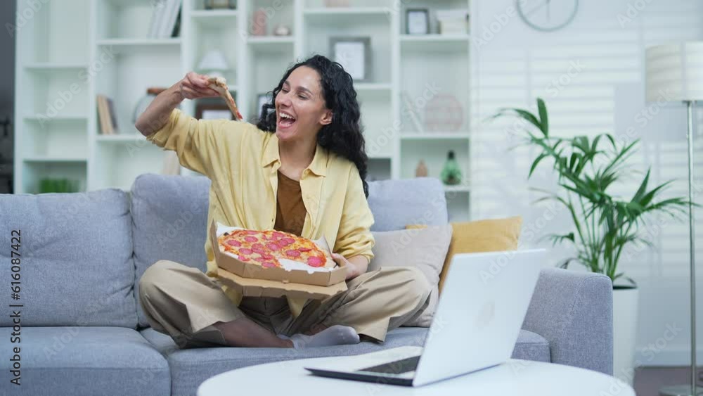 Young cheerful woman eating pizza sitting on sofa in living room. A smiling female holds a paper box in her hands and is happy about the fast delivery of food. The student ordered food to take home Young cheerful woman eating pizza sitting on sofa in living room. A smiling female holds a paper box in her hands and is happy about the fast delivery of food. The student ordered food to take home