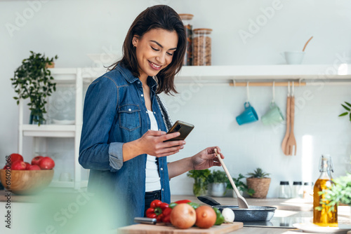 Obraz na plátně Smiling beautiful woman using her mobile phone while cooking vegetables in the kitchen at home