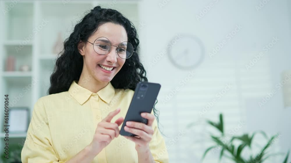 Close up. Happy young brunette woman using smartphone in living room at home. Smiling female in glasses browses social networks, makes purchases in online store, chats with friends, writes messages Close up. Happy young brunette woman using smartphone in living room at home. Smiling female in glasses browses social networks, makes purchases in online store, chats with friends, writes messages