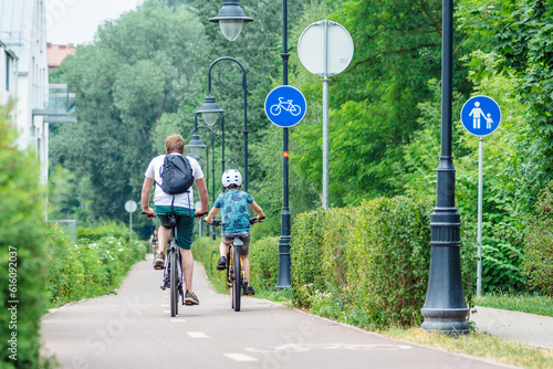 Father with backpack and son with helmet cycling together with bikes on the bike road with blue road sign or signal of bicycle lane among green trees and hedges, spring summer nature background