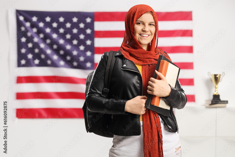 Female student with a hijab holding books and looking at camera in front of USA flag