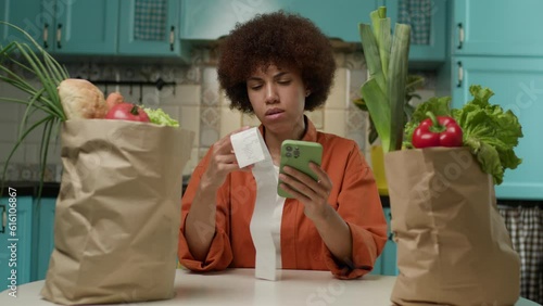 Black Woman Checking Shopping List with Receipt and Smartphone. 20s female looking at grocery bags full of fresh food and mobile phone. photo