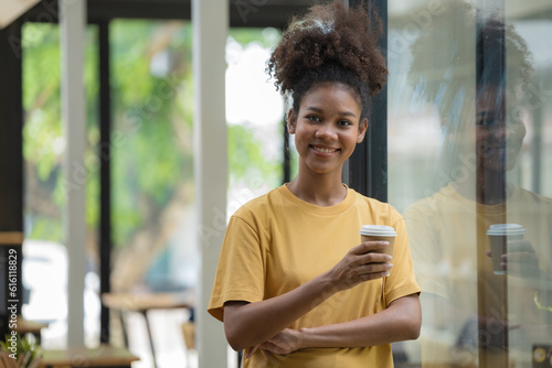 A smiling black woman drinking coffee while standing near a window at a cafe, a Happy young African American female holding a cup with a hot drink and looking at the camera.