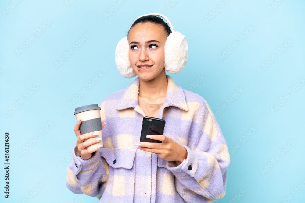 Young Arab woman wearing winter muffs isolated on blue background holding coffee to take away and a mobile while thinking something Young Arab woman wearing winter muffs isolated on blue background holding coffee to take away and a mobile while thinking something