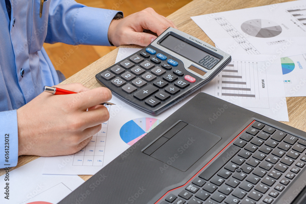 Mężczyzna siedzący przy biurku w pracy, liczący na kalkulatorze , szykujący ofertę dla klienta  - obrazy, fototapety, plakaty 