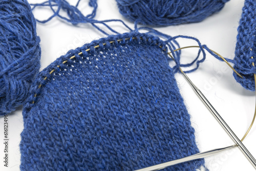 Blue wool yarn knitting on a metal needle with yarn balls on a white background, stockinette stitch knitted texture in progress
