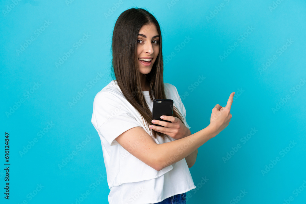 Young caucasian woman isolated on blue background using mobile phone and pointing back Young caucasian woman isolated on blue background using mobile phone and pointing back