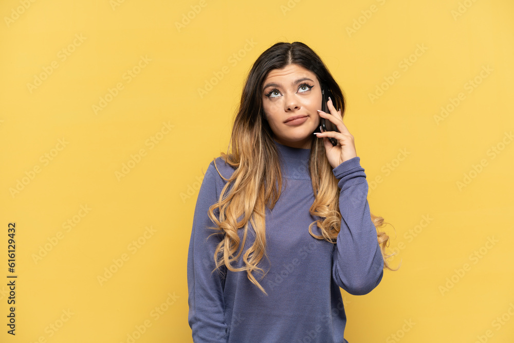 Young Russian girl using mobile phone isolated on yellow background and looking up Young Russian girl using mobile phone isolated on yellow background and looking up