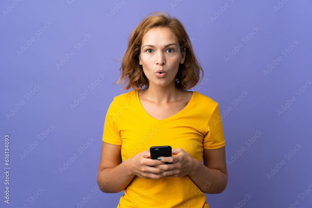 Young Georgian woman isolated on purple background looking at the camera while using the mobile with surprised expression Young Georgian woman isolated on purple background looking at the camera while using the mobile with surprised expression