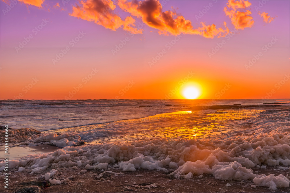 Sun setting on the horizon over the sea and the beach covered with golden foam.