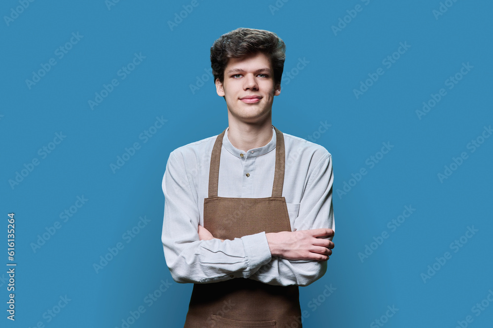 Fototapeta premium Portrait of young guy in apron with crossed arms on blue background