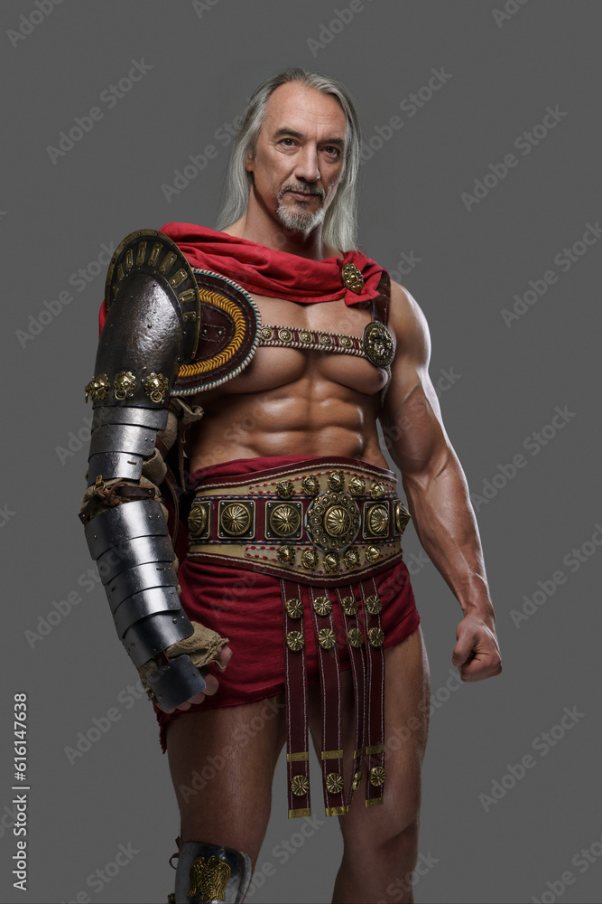 Mature and muscular gladiator with a stylish grey beard and flowing silver locks dons lightweight armor posing against grey backdrop Mature and muscular gladiator with a stylish grey beard and flowing silver locks dons lightweight armor posing against grey backdrop