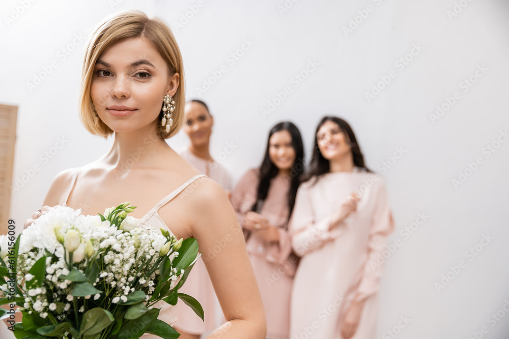 attractive bride in wedding dress holding bridal bouquet, standing near blurred interracial bridesmaids on grey background, happiness, special occasion, blonde and brunette women attractive bride in wedding dress holding bridal bouquet, standing near blurred interracial bridesmaids on grey background, happiness, special occasion, blonde and brunette women