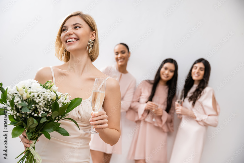 cheerful bride in wedding dress holding bridal bouquet and champagne glass, standing near blurred interracial bridesmaids on grey background, happiness, special occasion, blonde and brunette women cheerful bride in wedding dress holding bridal bouquet and champagne glass, standing near blurred interracial bridesmaids on grey background, happiness, special occasion, blonde and brunette women