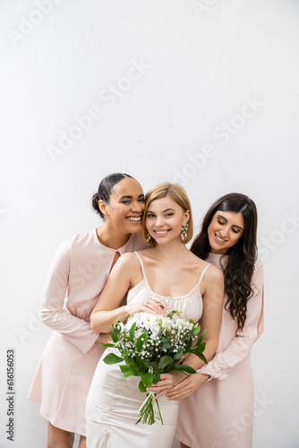 bridal party, special occasion, brunette and interracial bridesmaids hugging blonde bride, friendship goals, grey background, cheerful multicultural girlfriends, cultural diversity, bridal bouquet