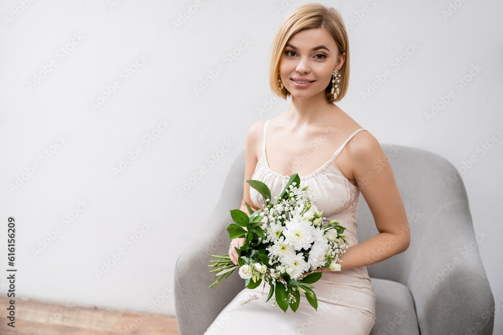 special occasion, beautiful blonde bride in wedding dress sitting in armchair and holding bouquet on grey background, looking at camera, white flowers, bridal accessories, happiness, feminine special occasion, beautiful blonde bride in wedding dress sitting in armchair and holding bouquet on grey background, looking at camera, white flowers, bridal accessories, happiness, feminine