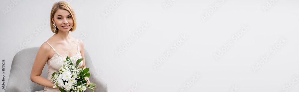 special occasion, beautiful, blonde bride in wedding dress sitting in armchair and holding bouquet on grey background, white flowers, bridal accessories, happiness, banner special occasion, beautiful, blonde bride in wedding dress sitting in armchair and holding bouquet on grey background, white flowers, bridal accessories, happiness, banner