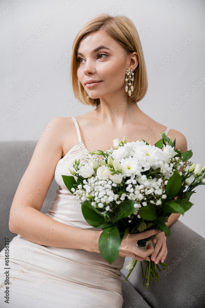 special occasion, beautiful and blonde bride in wedding dress sitting in armchair and holding bouquet on grey background, white flowers, bridal accessories, feminine, blissful special occasion, beautiful and blonde bride in wedding dress sitting in armchair and holding bouquet on grey background, white flowers, bridal accessories, feminine, blissful