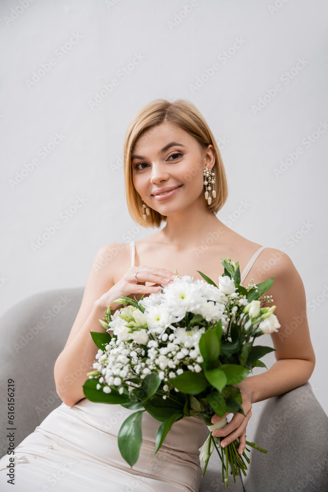 special occasion, beautiful blonde bride in wedding dress sitting in armchair and holding bouquet on grey background, engagement ring, white flowers, bridal accessories, happiness, feminine