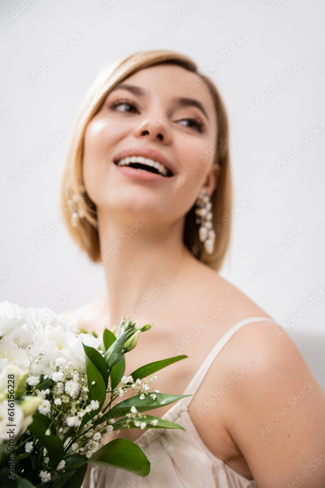 special occasion, beautiful, blonde bride in wedding dress holding bouquet on grey background, white flowers, bridal accessories, happiness, feminine, blissful, looking away, joy